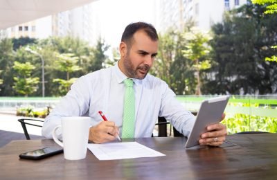 Serious mature freelancer working with digital tablet at cafe. Portrait of Caucasian businessman sitting at table and communicating to partner via pc tablet. Freelance concept